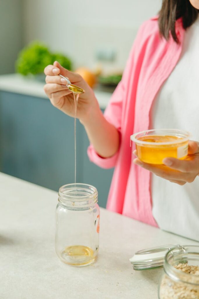 Close-up of a woman pouring honey from a spoon into a glass jar in the kitchen, healthy lifestyle